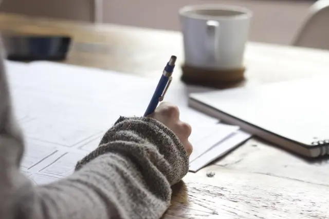 A hand writing in a notebook on a desk beside a cup of coffee