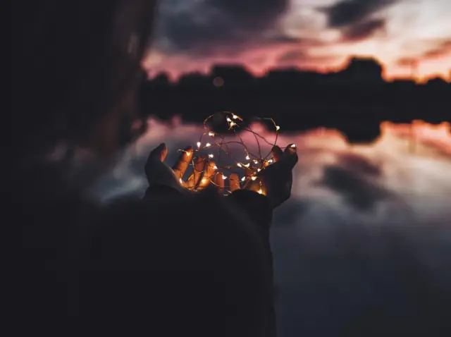 Hands holding a string of small glowing lights at dusk by the water