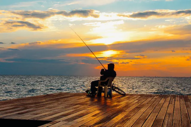 The silhouette of a man fishing from a wooden dock at sunset over the water